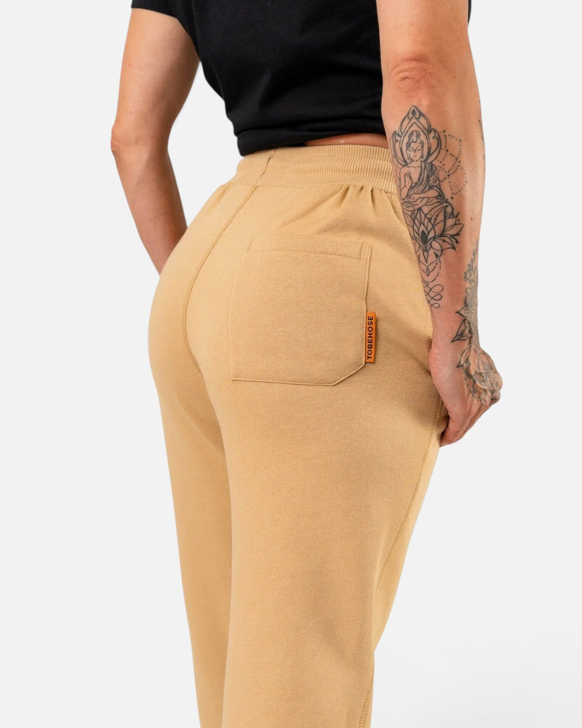 Tobe Hose Studio Damen Jogginghose in Beige mit Gesäßtasche und Markenlabel, getragen von einer Frau mit Tattoo