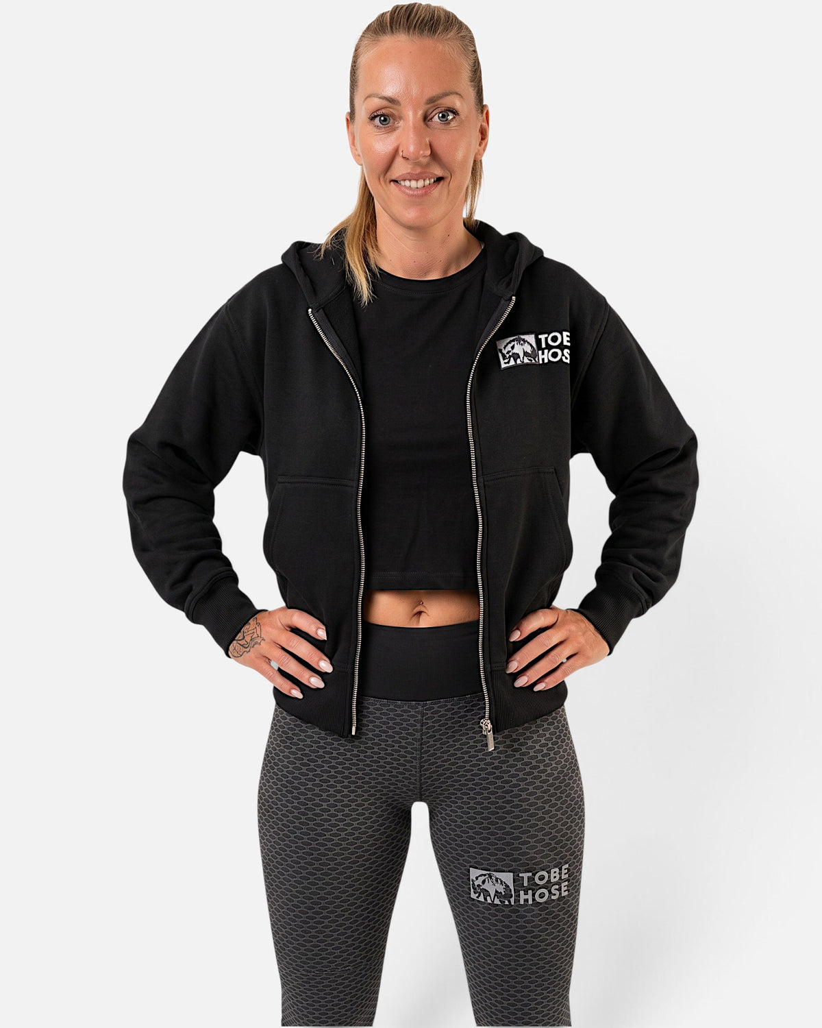 Tobe Hose Studio Damen Zip-Hoodie Fels in Schwarz mit Hyänen-Logo auf der Brust, getragen von einer Frau.