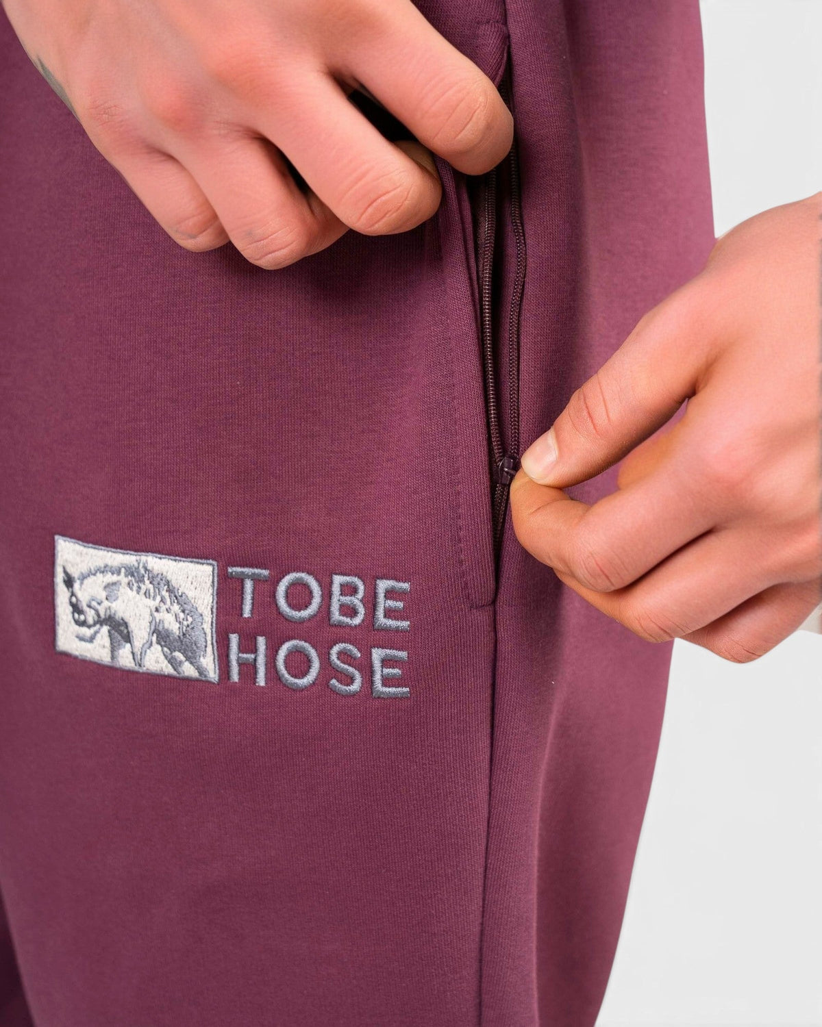 Tobe Hose Studio Herrenhose in Pflaume mit Reißverschlusstasche und gesticktem Hyänen-Logo auf robustem Stoff