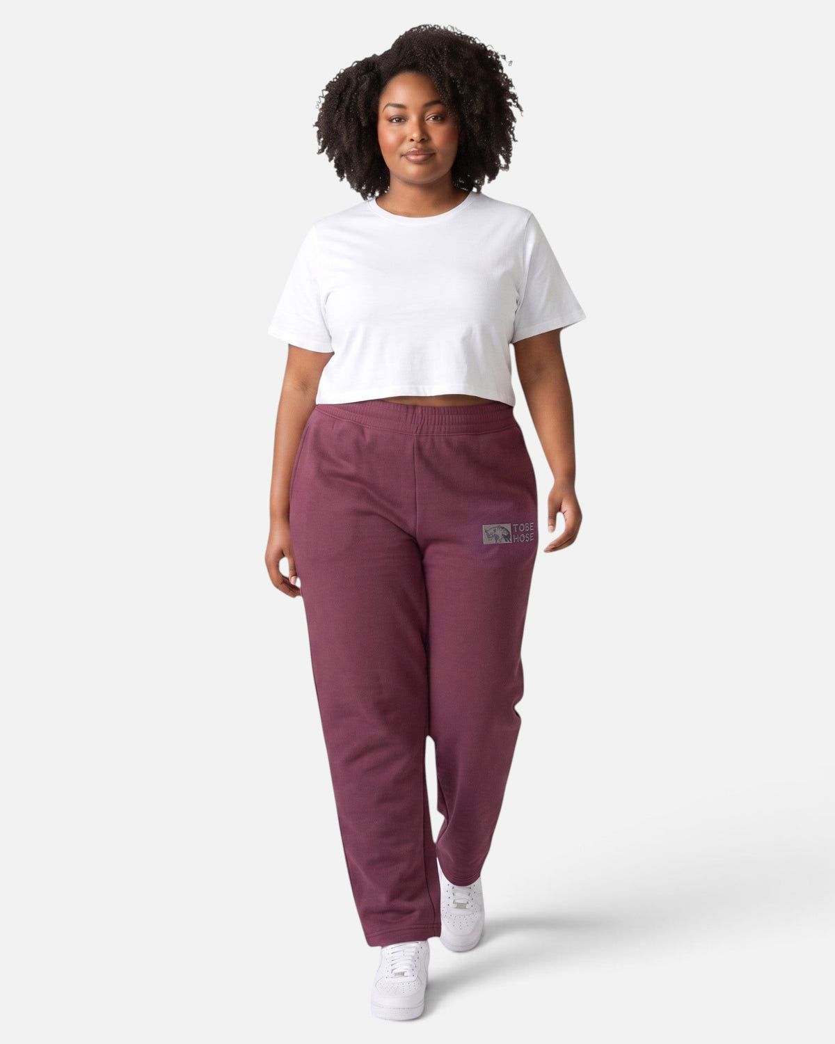 Model trägt Tobe Hose Studio Jogginghose in Pflaume mit Hyänen-Logo-Stick und Seitentaschen, kombiniert mit weißem Top.