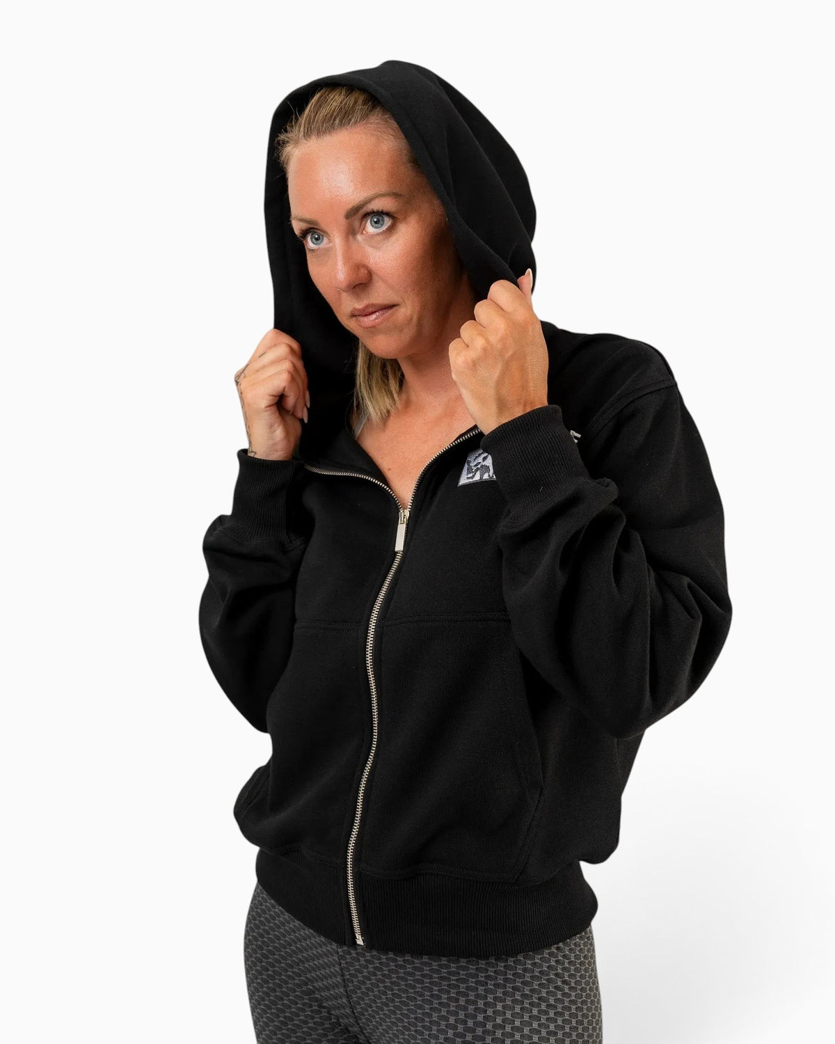 Tobe Hose Studio Zip-Hoodie Fels in Schwarz mit Hyänenmotiv auf der Brust, getragen von einer Frau mit Kapuze